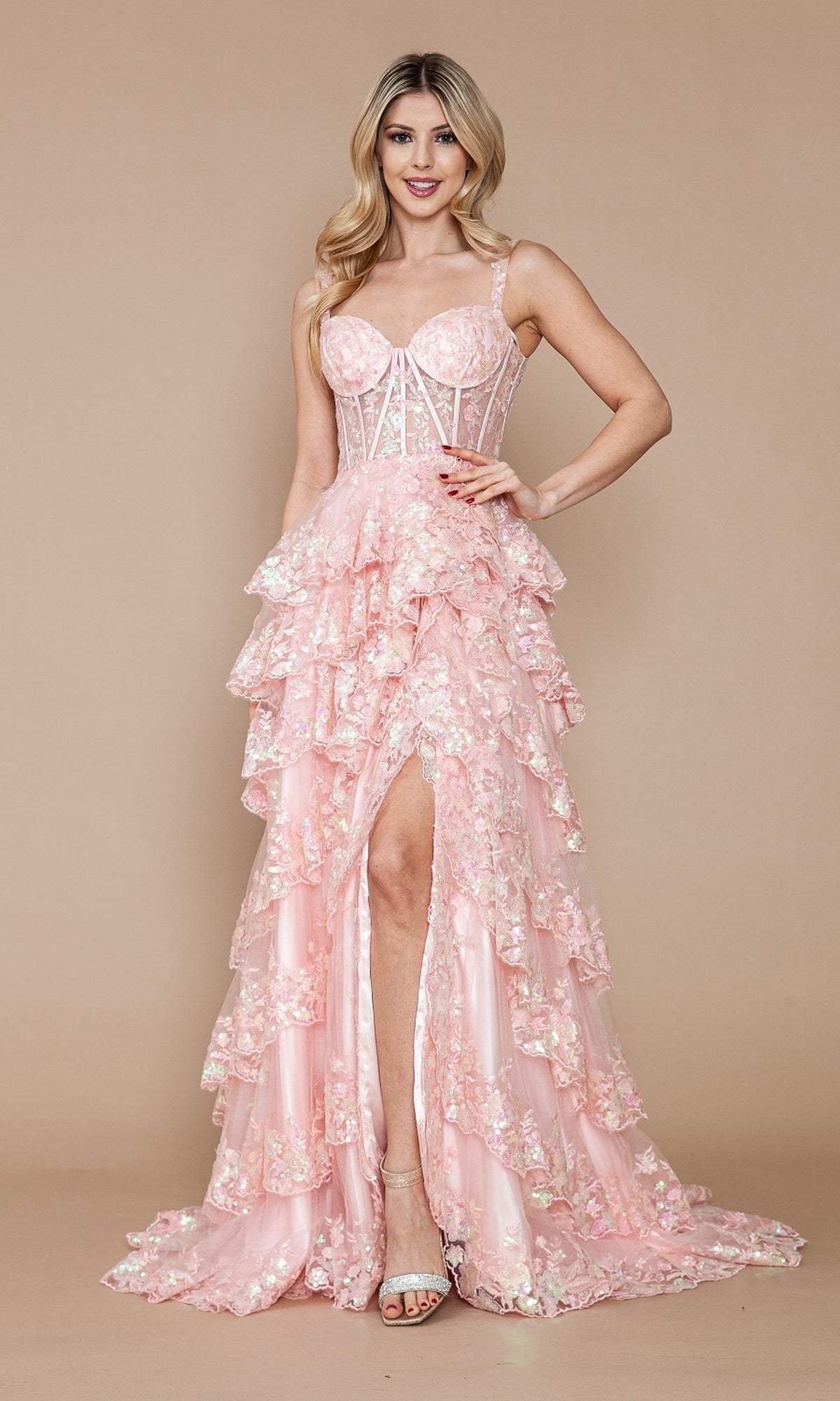 A woman wears the Sheer-Corset Long Ruffled Prom Dress (Poly USA 9410), featuring a pink, tiered floral lace skirt, corset bodice, and front slit, standing against a beige background.